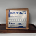 A package of Parea Coffee Co. Natural Guatemala Atitlan Pomegranate and Grapefruit Black Tea stands on a dark wooden tray, with a small pile of brewed dark tea on the white surface below.