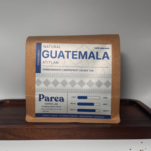 A package of Parea Coffee Co. Natural Guatemala Atitlan Pomegranate and Grapefruit Black Tea stands on a dark wooden tray, with a small pile of brewed dark tea on the white surface below.